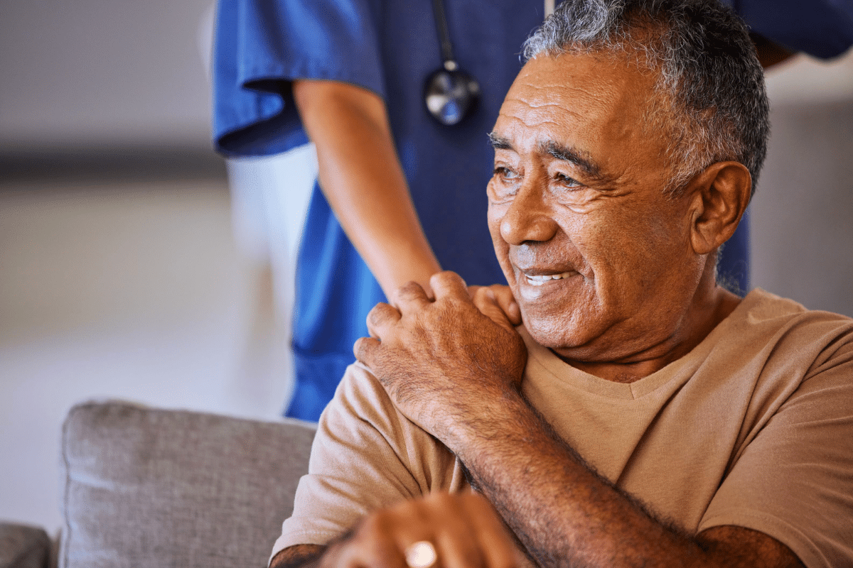 older adult with a cane sits on the couch with a nurse comforting them from behind
