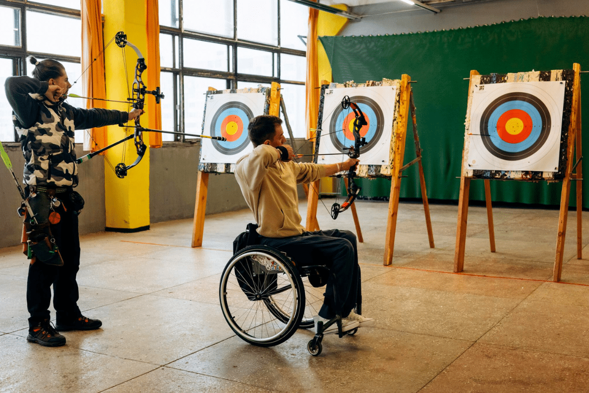 a young adult in a wheelchair plays archery along with a standing friend