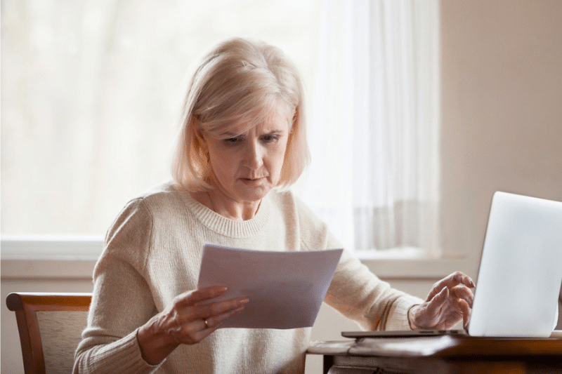 a concerned older adult woman reading paperwork at her opn laptop